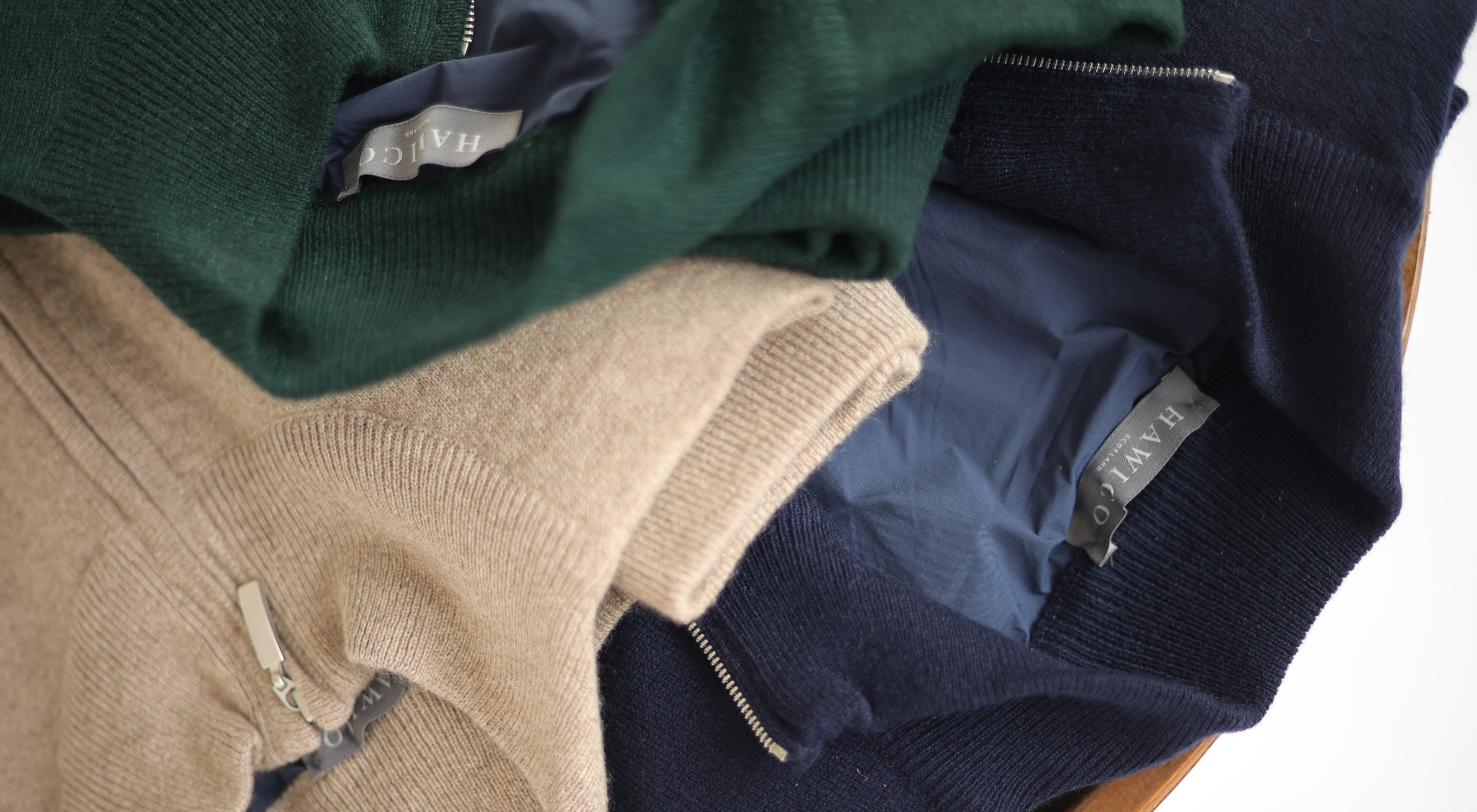 Stack of folded sweaters in green, beige, and navy on a wooden surface.